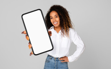 A cheerful young black woman with curly hair is holding a smartphone with a blank screen towards...