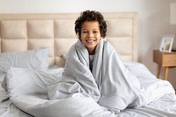 African American child is sitting on a bed, wrapped snugly in a soft blanket. The child eyes are...