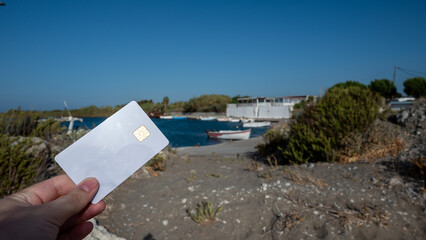 A woman's hand holds a white bank card against a background of a small cove with fishing boats.