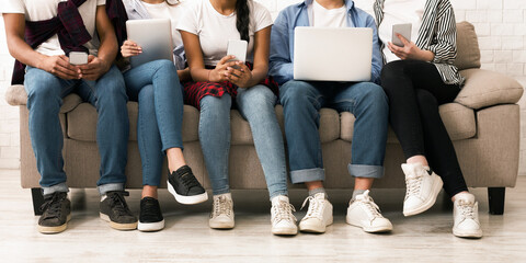 Cropped of five multiethnic teenagers seated side by side on a comfortable beige sofa, each engaged with different digital devices including smartphones, tablets, and laptops