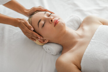 A serene young woman is lying down, eyes closed, experiencing the soothing touch of a head massage...