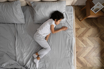 A young African American child lies curled up on a white bed, in a peaceful slumber, with gentle sunlight filtering through the room, casting soft shadows on the wooden floor, top view