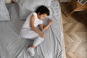 African American young child with curly hair is taking a nap, curled up on a neatly made grey bed...