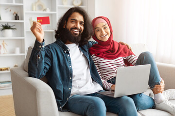 Happy young Indian couple sitting on couch together, using credit card and computer laptop, banking...