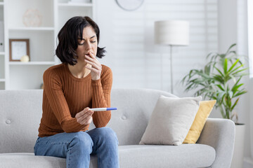An upset young woman is sitting on the sofa at home and looking disappointedly at the fake pregnancy test she is holding in her hands