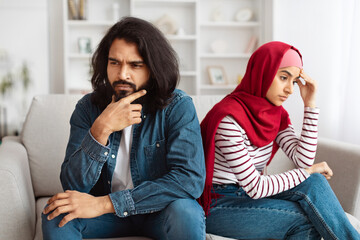 A bearded Indian man and a woman in a red hijab are sitting separately on a couch, showing...