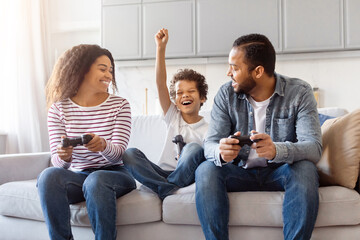 A joyful African American family is gathered on a cozy sofa in living room, engaged in gaming...