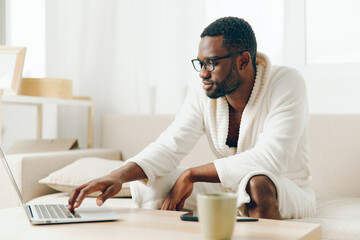 Smiling African American Freelancer Working on Laptop in Bathrobe on Sofa in the Morning This...