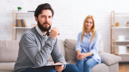 A bearded man therapist, holding a notebook, is seated attentively with a contemplative expression,...