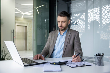 Fototapeta premium A professional male in a smart suit works attentively at his laptop in a well-lit contemporary office space, showing dedication and productivity.