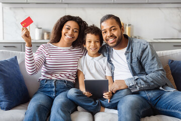 A joyful African American family is gathered on the sofa in a cozy, well-lit living room, using...