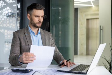 Focused professional male in a suit analyzing papers with a laptop in a contemporary office environment. Represents work, concentration, and corporate lifestyle. © Liubomir