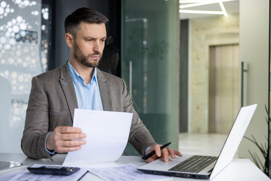 Focused professional male in a suit analyzing papers with a laptop in a contemporary office environment. Represents work, concentration, and corporate lifestyle.