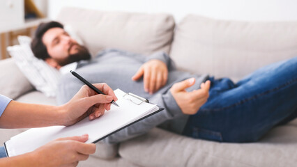 Stressed man telling about his problems to doctor, lying on couch at psychotherapist office, doctor making notes