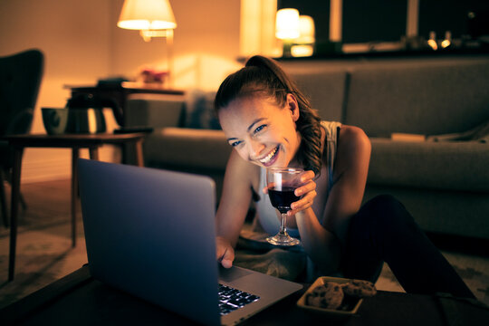 Relaxed young woman with wine using laptop at home in the evening