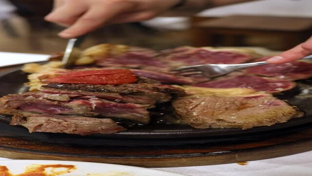 Process of cutting the medium rare steak with vegetables on a cutting board. The juicy beef meat is cut into pieces with fork and knife. Gourmet menu at restaurant. Unrecognizable person eat beef