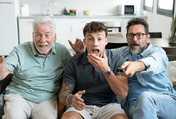 Males of multigenerational Caucasian family sitting together at home enthusiastically cheering for the athletes of the Olympic games. Sport and participation of three attractive men of different ages