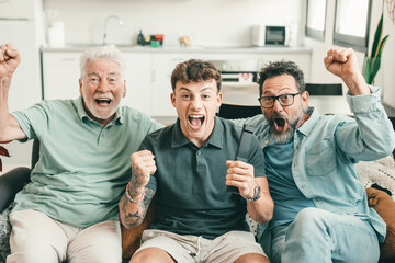 Males from multigenerational family sitting together at home looking at tv enthusiastically cheer athletes of Olympic games. Sports and participation of three attractive men of different ages