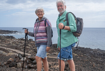 Smiling senior couple walks in a seaside path with help of a stick, elderly pensioners leads healthy life walking outdoors in Tenerife, Canary Islands. Horizon over water of atlantic ocean