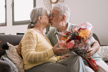 Senior man giving bouquet of flowers at his wife sitting on the sofa at home for anniversary or San Valentines’ day. Pensioners kissing enjoying surprise together. In love people having fun.