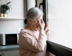 Pensive sad elderly mature senior woman with hand on eyes standing near window, thinking of personal problems