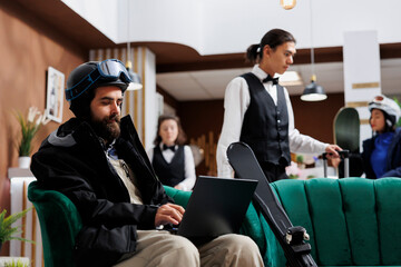 A caucasian male traveler seated at hotel lobby browsing on a laptop. Young man wearing a winter...