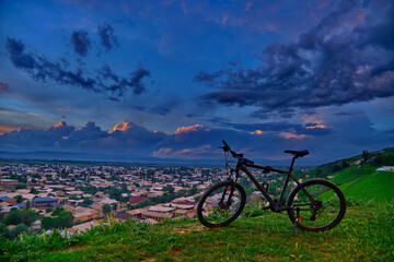 A mountain bike standing atop a green hill with a city panorama against a stormy sky in the...