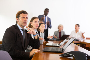 Fototapeta premium Middle-aged white brunette businessman furrowing his eyebrows and squinting thus expressing his objection and surprise while sitting at corporate meeting in conference room