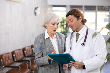 Adult man in medical uniform and elderly woman in business suit look at folder with documents in clinic reception