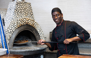 Smiling African American man chef preparing pizza in restaurant, taking pizza from oven at kitchen