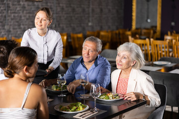 Adult woman waiter serving glass of wine to elderly couple and young couple in restaurant