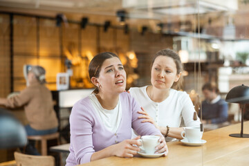 Middle-aged woman comforting her unhappy female friend while sitting with cups of coffee or tea in...