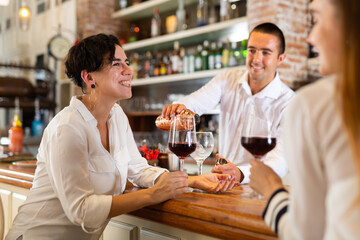 Two girlfriends chat with each other and drink red wine while sitting at bar in a restaurant