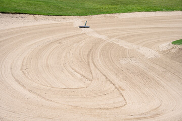 Closeup of large, groomed sand trap with rake on a golf course, tracks of previous golfer who’s...