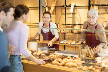 Portrait of two female bakery workers of different ages in uniform selling pastry to consumers in bakehouse