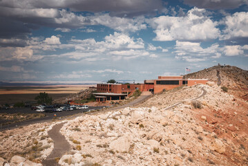 Landscape in Arizona, USA with rocky terrain leading to a building complex, likely near Meteor Crater. Expansive sky and American flag add to the atmosphere. Car park indicates popular destination. - Powered by Adobe