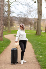 Woman in sunglasses stands with a suitcase on a park alley, facing the camera.