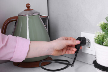 Woman plugging electric kettle into power socket at white table indoors, closeup