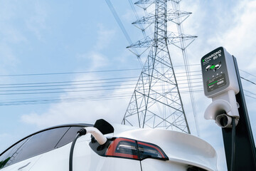 EV electric car at recharging at charging station connected to electrical power grid tower on sky...