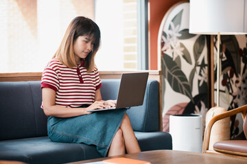 Fototapeta premium Concentrated young woman in a striped shirt working on a laptop while seated on a couch in a stylish cafe.
