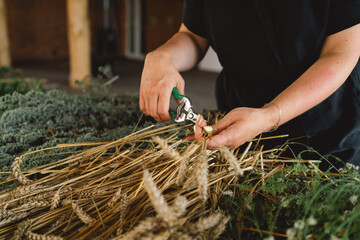 A woman stands at a wooden table in a rustic, diligently working on assembling a natural wreath from an assortment of freshly cut branches and foliage that are scattered across the work surface.
