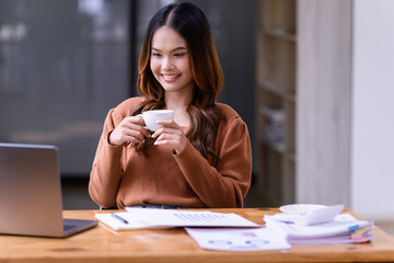 A young woman holding a cup of coffee relaxing sitting at working desk in the office.