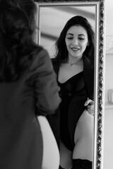 Seductive girl in black bodysuit, stockings and jacket. Smiling and admiring herself in the mirror. Black and white