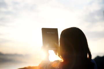 Christian woman holding high holy bible with sunset sky and bright light sun background, Christian...