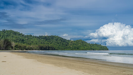 A serene seascape. The waves of the turquoise ocean roll towards the shore, foaming and spreading over the sandy beach. A hill covered with green tropical vegetation against a blue sky and clouds. 