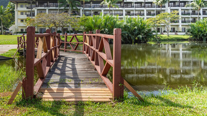 A wooden pedestrian bridge with a lattice railing spans over the stream. Lush tropical plants, palm trees on the shore. Reflection on calm water. The hotel building is visible in the distance.