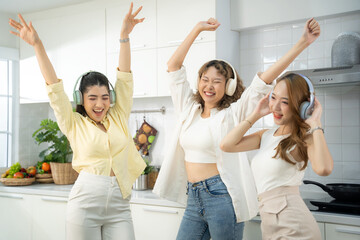 Three young friends dance joyfully in a bright kitchen, headphones on, expressing happiness and carefree lifestyle.