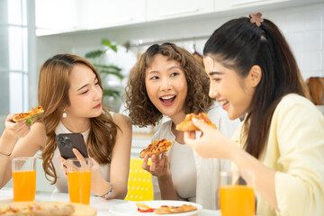 Three happy women enjoying pizza and orange drinks while chatting and using a smartphone in a modern kitchen.