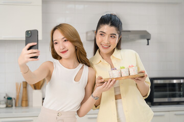 Two smiling women taking a selfie in a modern kitchen, showcasing a wooden tray with homemade decorated cupcakes.