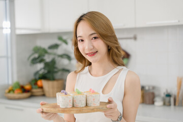 Smiling woman holding a wooden tray with decorated homemade cupcakes, standing in a modern kitchen.